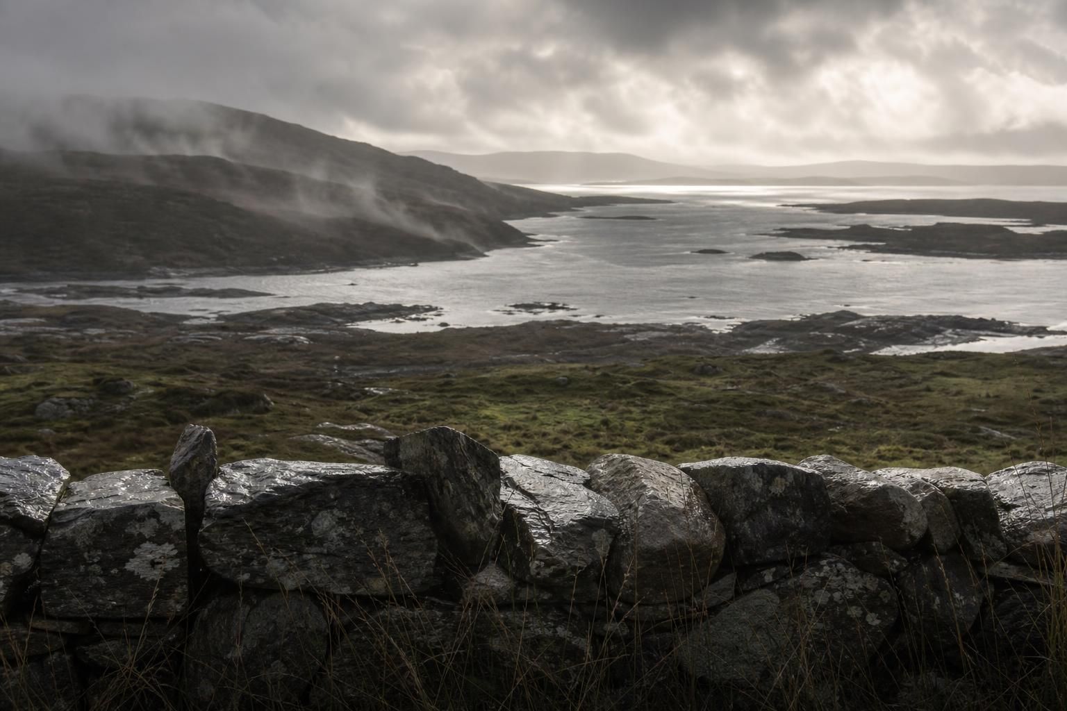 After-rain Connemara coastline on Ireland's Atlantic edge: wet stone wall, peat moor, mist and cloud movement over a silver inlet in soft Atlantic light at dusk